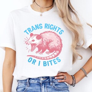 May include: A white short-sleeve t-shirt featuring a pink illustration of an opossum in the center. The text "TRANS RIGHTS OR I BITES" is printed in light blue capital letters in an arc above and below the opossum graphic.