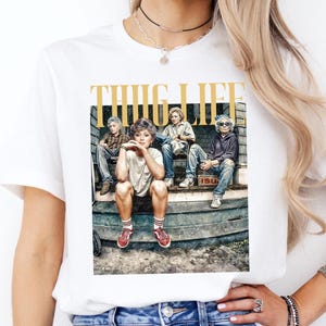 May include: White t-shirt with a graphic print of four people sitting on a porch. The graphic has the text "THUG LIFE" in gold lettering.