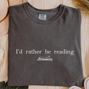 May include: Dark gray t-shirt with the text "I'd rather be reading" and a small book graphic. The shirt is surrounded by a beige hat, white headband, white sneakers, and decorative elements.