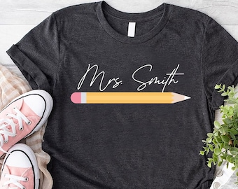 May include: A dark gray t-shirt with the text "Mrs. Smith" in white cursive writing and a yellow pencil with a pink eraser above the text.