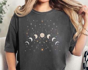 May include: A woman wearing a dark gray t-shirt with a moon and stars design. The shirt has a distressed look and is a relaxed fit.