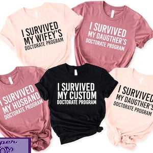 May include: Five t-shirts with different text designs. The shirts are in various colors, including pink, black, and peach. The text on the shirts reads: "I survived my wifey's doctorate program", "I survived my daughter's doctorate program", "I survived my husband's doctorate program", "I survived my custom doctorate program", and "I survived my daughter's doctorate program".