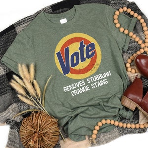 May include: Green t-shirt with a vintage-style logo that says "Vote" in a blue circle with a red and yellow background. The text below the logo says "Removes Stubborn Orange Stains".