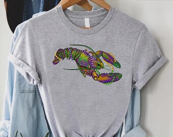 May include: A gray t-shirt with a purple and green lobster wearing Mardi Gras beads. The lobster is facing the right side of the image.