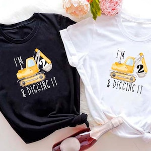 May include: Two short-sleeved t-shirts, one black and one white, each featuring a yellow excavator graphic with the text "I'M & DIGGING IT" and the number "2". The shirts are likely for a second birthday celebration.