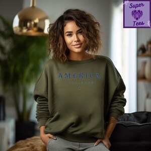 Puede incluir: Sudadera verde oliva con la palabra "AMERICA" en letras azules en el pecho. Debajo, el texto "1776-2020" y una peque&ntilde;a estrella azul. La sudadera es de cuello redondo.