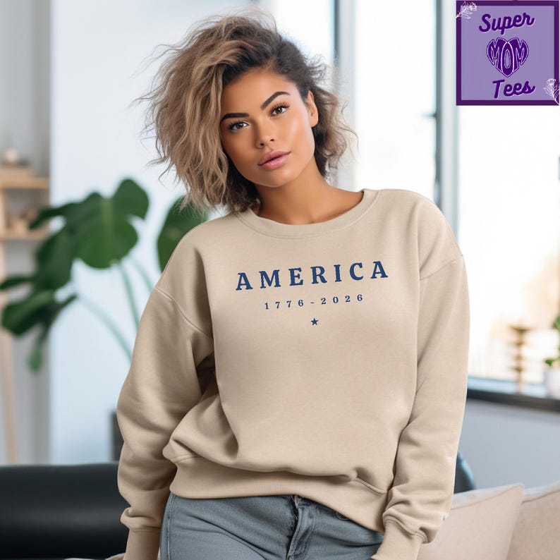 Puede incluir: Una sudadera beige de cuello redondo con la palabra "AMERICA" en letras azules oscuras, junto con las fechas "1776-2026" y una peque&ntilde;a estrella. La sudadera es de color neutro y tiene un estilo cl&aacute;sico e informal.