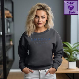 Puede incluir: Sudadera gris oscuro con la palabra "AMERICA" en letras azules en el pecho. Debajo, los n&uacute;meros "1776-2023" y una peque&ntilde;a estrella azul. La sudadera forma parte de la colecci&oacute;n Super Mom Tees.