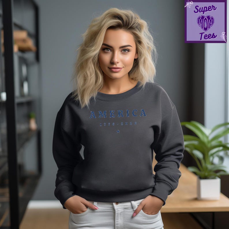 Puede incluir: Sudadera gris oscuro con la palabra "AMERICA" en letras azules en el pecho. Debajo, los n&uacute;meros "1776-2023" y una peque&ntilde;a estrella azul. La sudadera forma parte de la colecci&oacute;n Super Mom Tees.
