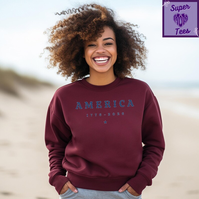 Puede incluir: Sudadera burdeos con la palabra "AMERICA" en letras azules. Debajo de la palabra "AMERICA" est&aacute; el texto "1776-2020" y una estrella. La sudadera es de cuello redondo.