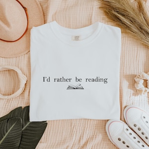 May include: White t-shirt with the text "I'd rather be reading" and an illustration of an open book. The shirt is surrounded by a beige hat, a white headband, a scrunchie, a pair of white sneakers, and green leaves.
