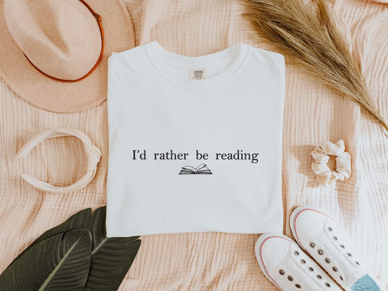 May include: White t-shirt with the text "I'd rather be reading" and an illustration of an open book. The shirt is surrounded by a beige hat, a white headband, a scrunchie, a pair of white sneakers, and green leaves.