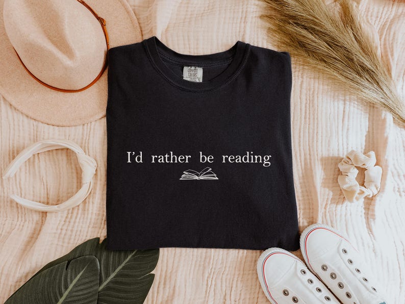 May include: Black t-shirt with the text "I'd rather be reading" and an open book graphic. The shirt is surrounded by a beige hat, white headband, scrunchie, green leaves, and white sneakers.