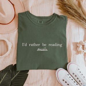 May include: An olive green t-shirt with the text "I'd rather be reading" and an open book graphic. The shirt is laid flat with a hat, headband, shoes, and decorative elements.