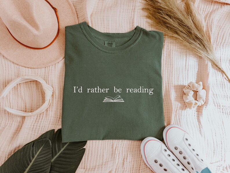 May include: An olive green t-shirt with the text "I'd rather be reading" and an open book graphic. The shirt is laid flat with a hat, headband, shoes, and decorative elements.