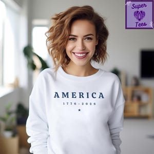 Puede incluir: Sudadera blanca con la palabra "AMERICA" en azul marino, las fechas "1776 - 2026" y una estrella. La persona que lleva la sudadera tiene el pelo casta&ntilde;o y est&aacute; sonriendo.