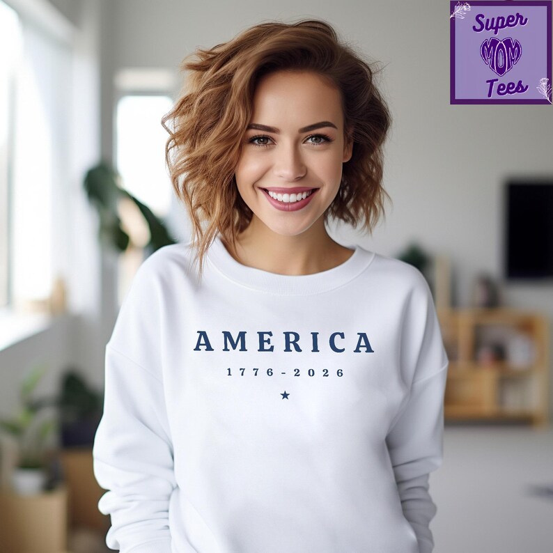 Puede incluir: Sudadera blanca con la palabra "AMERICA" en azul marino, las fechas "1776 - 2026" y una estrella. La persona que lleva la sudadera tiene el pelo casta&ntilde;o y est&aacute; sonriendo.