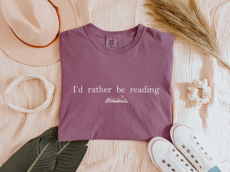 May include: A mauve t-shirt with the text "I'd rather be reading" and an open book graphic. The shirt is surrounded by a cream-colored hat, headband, scrunchie, and white sneakers.