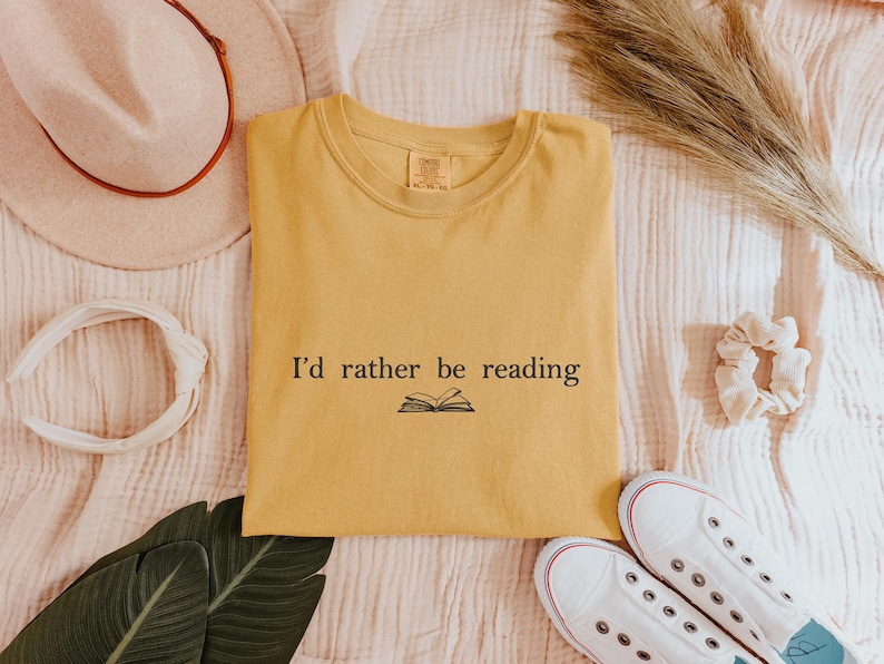 May include: Mustard yellow t-shirt with the text "I'd rather be reading" and an open book graphic. The shirt is surrounded by a hat, headband, scrunchie, sneakers, and decorative leaves, all on a textured surface.