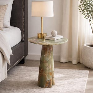 May include: A green and brown marble side table with a round top and a pedestal base. A gold-toned lamp with a white shade sits on the table, along with a small, round, white decorative object and a book. The table is placed on a light-colored rug.