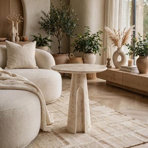 May include: A round, beige travertine side table with a textured, porous surface and a tapered pedestal base, positioned on a light-colored area rug in front of a curved, cream-colored sofa with a matching throw blanket and pillow.