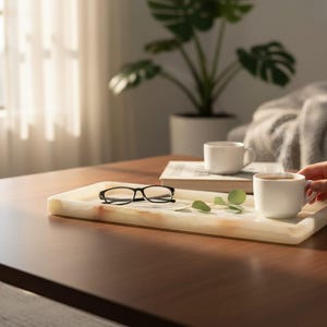 May include: A rectangular white and cream-colored stone tray with natural veining. Resting on the tray are a pair of black-rimmed eyeglasses and a small green sprig. A white ceramic mug sits on the edge of the tray.