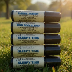 May include: A stack of five essential oil roll-on bottles with black caps and cream-colored labels. The labels read "HAPPY TUMMY", "BOO BOO BLEND", and "SLEEPY TIME". The bottles are on a bed of green grass.