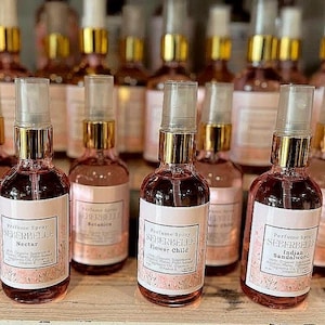 May include: A collection of perfume spray bottles with gold-colored tops and clear spray nozzles. Each bottle has a pink liquid and a white label with the brand name "SEBERBELLE" and a fragrance name. The bottles are arranged in rows.