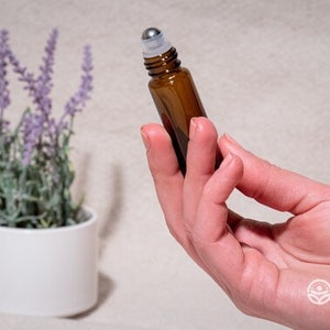 Op de afbeelding: Een bruine glazen rollerfles met een zilveren metalen rollerbal. De fles wordt in een hand gehouden.