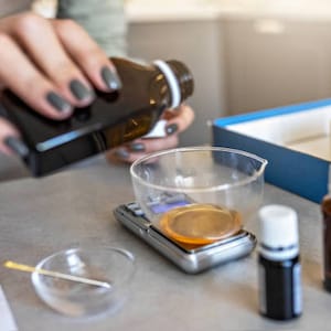Op de afbeelding: Een persoon giet vloeistof uit een donkerbruine fles in een glazen maatbeker op een digitale weegschaal. Er staan twee kleine flesjes met donkere vloeistof op een wit oppervlak.