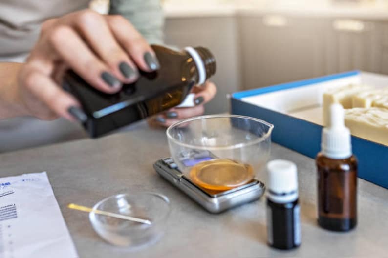 Op de afbeelding: Een persoon giet vloeistof uit een donkerbruine fles in een glazen maatbeker op een digitale weegschaal. Er staan twee kleine flesjes met donkere vloeistof op een wit oppervlak.