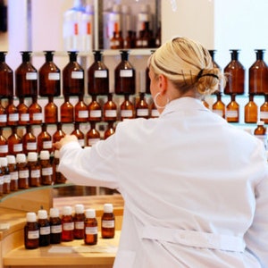 Op de afbeelding: Een vrouw in een witte labjas staat voor planken vol met rijen amberkleurige glazen flessen. De flessen zijn in een rasterpatroon gerangschikt en bevatten verschillende vloeistoffen.