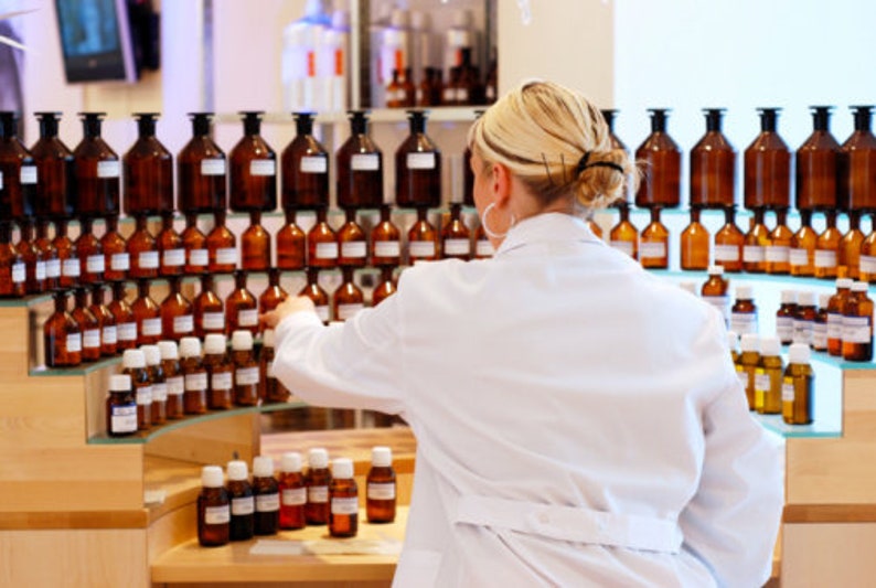Op de afbeelding: Een vrouw in een witte labjas staat voor planken vol met rijen amberkleurige glazen flessen. De flessen zijn in een rasterpatroon gerangschikt en bevatten verschillende vloeistoffen.