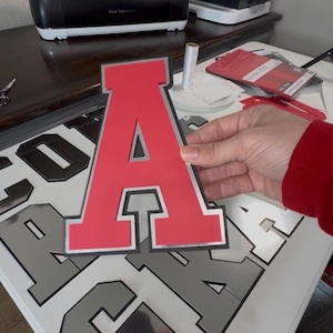 Puede incluir: Se muestra una letra "A" roja con borde plateado. Se ven otras letras sobre una superficie blanca. La letra forma parte de un conjunto, posiblemente para un letrero o decoración. Suministros de manualidades en el fondo.