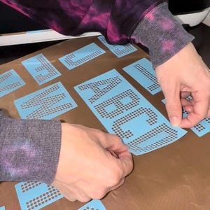 May include: A set of light blue adhesive stencils featuring individual letters of the alphabet formed by small circular cutouts. The stencils are arranged on a brown surface, with the letters A, B, C, and D clearly visible on one sheet.