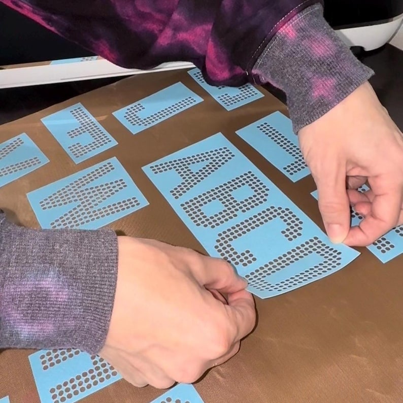 May include: A set of light blue adhesive stencils featuring individual letters of the alphabet formed by small circular cutouts. The stencils are arranged on a brown surface, with the letters A, B, C, and D clearly visible on one sheet.