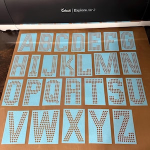 May include: A collection of blue letter stencils, each with a dotted pattern, arranged on a brown surface. The letters A through Z are visible, with the Cricut Explore Air 2 machine in the background.
