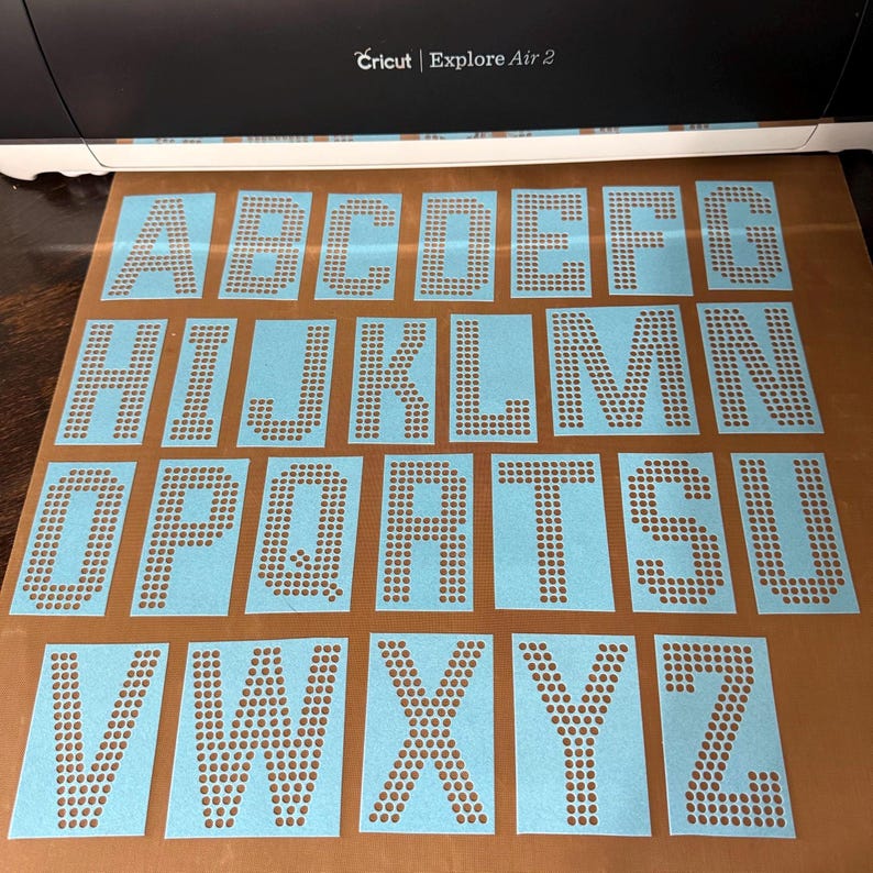 May include: A set of light blue adhesive alphabet stencils featuring the letters A through Z, each formed by a grid of small circular cutouts. The stencils are arranged in rows on a brown backing sheet, positioned in front of a Cricut Explore Air 2.
