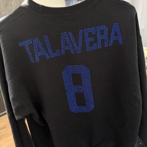May include: A black long-sleeve sweatshirt featuring the name TALAVERA and the number 8 on the back, both rendered in blue rhinestones. The garment has a crew neckline and long sleeves, with the text and number centered on the upper back area.