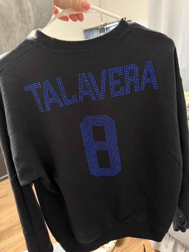 May include: A black long-sleeve sweatshirt featuring the name TALAVERA and the number 8 on the back, both rendered in blue rhinestones. The garment has a crew neckline and long sleeves, with the text and number centered on the upper back area.
