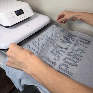 May include: A white heat press machine with a digital display showing 305 degrees and 15 seconds, positioned over a light gray t-shirt featuring a transfer sheet with the alphabet letters A through U arranged in rows, created with silver rhinestones.