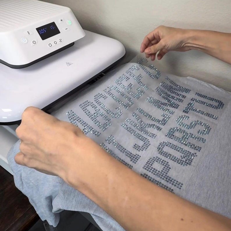 May include: A white heat press machine with a digital display showing 305 degrees and 15 seconds, positioned over a light gray t-shirt featuring a transfer sheet with the alphabet letters A through U arranged in rows, created with silver rhinestones.