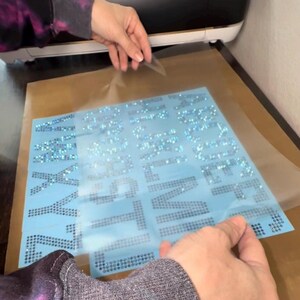 May include: A light blue transfer sheet featuring the alphabet in uppercase letters formed by small, reflective, iridescent rhinestones. The letters are arranged in columns on a clear adhesive backing, partially covered by a translucent protective film.