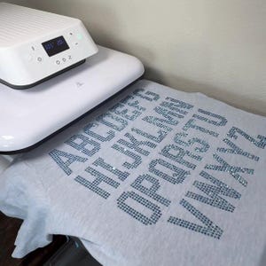 May include: A light gray t-shirt featuring the alphabet from A to Z spelled out in blue rhinestones. A white heat press machine with a digital display showing 305 degrees and 20 seconds is positioned over the top portion of the t-shirt.