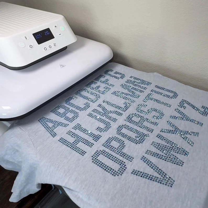 May include: A light gray t-shirt featuring the alphabet from A to Z spelled out in blue rhinestones. A white heat press machine with a digital display showing 305 degrees and 20 seconds is positioned over the top portion of the t-shirt.