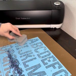 May include: A black and white Cricut Explore Air 2 machine sits behind a light blue adhesive sheet covered in small, dark blue rhinestones arranged to form the letters of the alphabet. A gray applicator tool is used to press the stones.