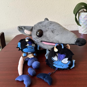 May include: A large grey plush rat with a black nose and large black eyes, accompanied by two smaller plush figures. The smaller figures have blue skin, black hair, and light-coloured antennae, with one featuring a long tail and the other a round head.