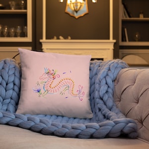 May include: A pink cushion featuring a colourful dragon design. The dragon is outlined in red and has a blue tail and green wings. The dragon is surrounded by pink and white flowers.