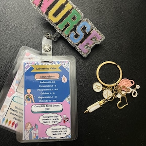 Puede incluir: Un llavero colorido 'NURSE' con purpurina y un anillo de llave dorado. El llavero incluye un pequeño amuleto de jeringa, un amuleto de corazón, un amuleto de mascarilla facial rosa y un amuleto con la letra 'N'. Un portagafete de plástico transparente con fondo blanco y borde azul muestra un cuadro de valores de laboratorio médico con los títulos 'Electrolitos' y 'Recuento sanguíneo completo CBC'.