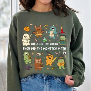 Könnte beinhalten: Grünes Sweatshirt mit einer bunten Grafik mit Cartoon-Monstern und dem Text "THEY DID THE MATH" und "THEY DID THE MONSTER MATH". Das Design umfasst einen Kürbis, eine Fledermaus, einen Geist und andere Halloween-Figuren.